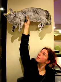 Barbarella hält eine handliche Version der Katze aus der Installation " Sing it again " in die Höhe; Barbarella blickt hinauf zur Katze und die Katze wiederum fixiert etwas was uns hier nicht gezeigt wird.